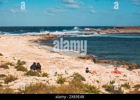 Sunbathers relaxing and reading in the sun on the rocky shores of Qawra Point Beach, St. Paul's Bay, Malta Stock Photo