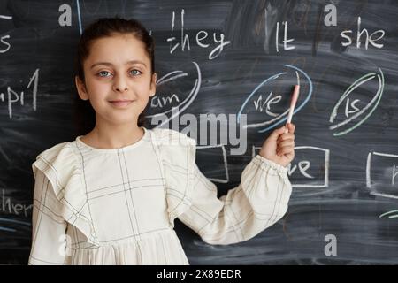 Portrait of Middle Eastern schoolgirl standing against blackboard in classroom showing English pronoun written on it and looking at camera Stock Photo
