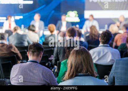 Diverse group of business professionals attending a conference, focusing on a panel discussion. The image captures the audience from behind, emphasizing active listening and engagement. Stock Photo