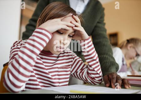 Medium closeup of Caucasian boy sitting at table in class struggling to do difficult task, unrecognizable teacher helping him Stock Photo