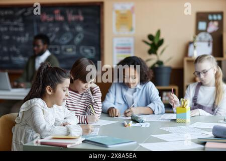 Group of four ethnically diverse boys and girls sitting at table in classroom doing exercises while their teacher using laptop in background Stock Photo