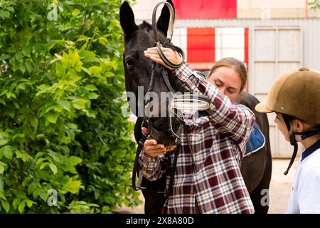 Woman instructor adjusting bridle on horse while interacting with young rider girl. Horse riding activity Stock Photo