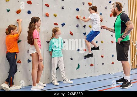 A diverse group of kids are energetically climbing and exploring a wall while being supervised by their male teacher in a bright, lively classroom set Stock Photo