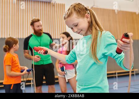 A diverse group of kids engages in an energetic gym session, with a girl confidently holding a jump rope as the male teacher instructs and guides them Stock Photo
