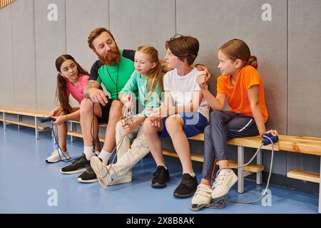 A group of diverse kids sit closely together on a bench, listening attentively to their male teacher in a vibrant gym. Stock Photo