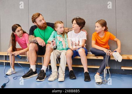A diverse group of children sitting attentively on a bench, listening to their male teacher in a bright, lively classroom setting. Stock Photo