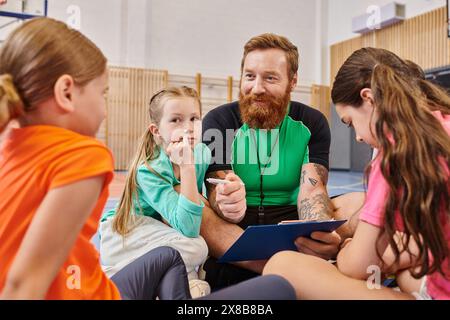 A bearded man sits on the basketball court surrounded by a diverse group of kids, mentoring and coaching them in an energetic and positive atmosphere. Stock Photo