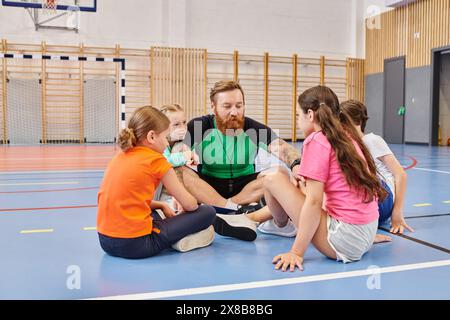 A male teacher sits on the floor surrounded by a diverse group of children in a vibrant school gym, engaged in interactive and educational activities. Stock Photo