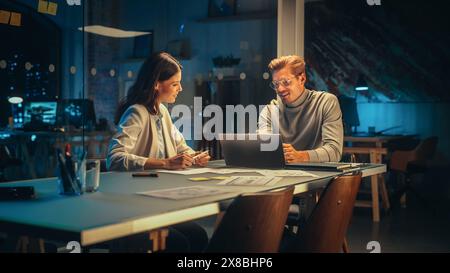 Small Creative Team Meeting in the Evening in Office. Young Project Manager and Excited Software Developer Using Laptop Computer, Discussing Updates to the New Online Application. Stock Photo