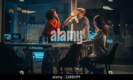 Small Creative Team Meeting in the Evening in Office. Young Project Manager Bringing Laptop Computer to Check and Discuss the Sales Number. Successful Managers High Five Each Other and Celebrate. Stock Photo