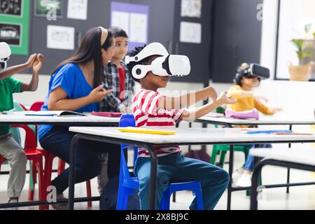 In school, diverse group of young students wearing virtual reality headsets in the classroom. A middle-aged biracial female teacher oversees activity, Stock Photo