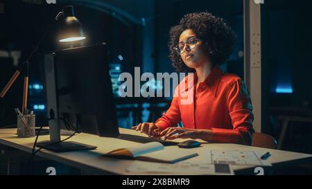 Young Black Female Working on Desktop Computer in Creative Office in the Evening. Happy Multiethnic Project Manager Writing Emails, Researching Project Plan Details Online. Stock Photo