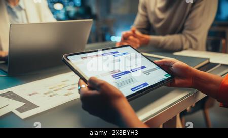 Positive Group of Managers Having a Team Meeting Long After Work Hours in the Night. Close Up on Tablet Computer Display with Work Management Software Stock Photo