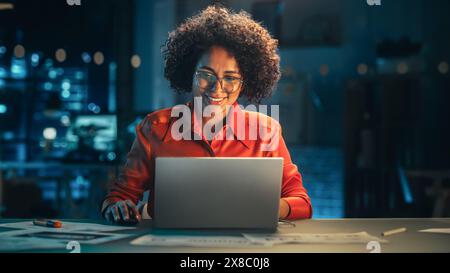 Young Black Female Working on Laptop Computer in Creative Office in the Evening. Happy Multiethnic Project Manager Writing Emails, Researching Project Plan Details Online. Stock Photo