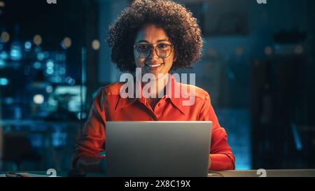 Beautiful Multiethnic Brazilian Female Working on Laptop Computer Late at Night. Young Creative Manager Feeling Happy, Looking at Camera and Smiling. Zoom In Portrait Footage. Stock Photo