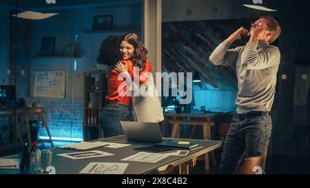 Excited Team Looking at Laptop Screen, Waiting for Business Partnership Tender Results Late in the Evening. Successful Managers Celebrating the Winning Result, High Five and Hug Each Other. Stock Photo