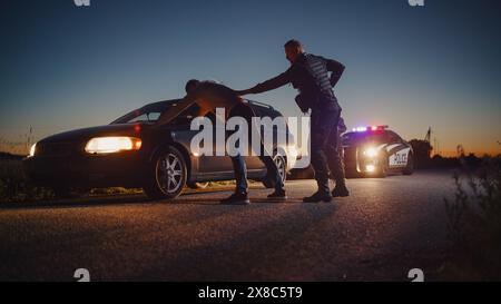 A Professional Middle Aged Policeman Performing a Pat-Down Search on a Fellon With his Hands on Car Hood. Documentary-like Shot of Procedure of Arresting Suspects. Experienced Cop Looking for Weapons Stock Photo