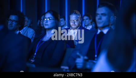 Young Bearded Man Attending an IT Conference in Crowded Audience. Delegate Using Laptop Computer. Happy Software Developer Watching Presentation About New High Tech Products and Data Protection. Stock Photo
