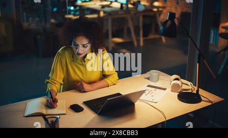Creative Multiethnic Female Working on Laptop Computer in a Company Office at Night. Happy Driven Project Manager Browsing Internet, Writing Tasks in Notebook, Developing a Marketing Strategy. Stock Photo