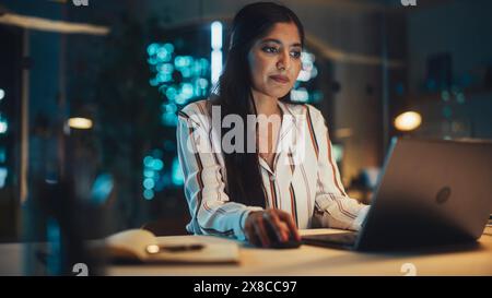 Portrait of a Beautiful Indian Female Working on Laptop Computer in a Company Office at Night. Young Secretary Analyzing Reports. Account Manager Developing a Marketing Strategy for a Client. Stock Photo