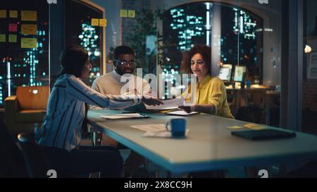 Small Diverse Multiethnic Group Collaborate on a Project and Work Together in an Office at Night. Team Leader Talking with a Professional, Discussing Changes to a Software Design. Stock Photo
