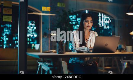 Beautiful Female in a Striped White Shirt Sitting at a Desk in Creative Office, Working on Tasks on Laptop Computer. Young Creative Specialist Writing Corporate Project Plan for Marketing Agenda. Stock Photo