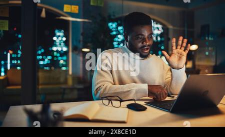 Young Handsome Black Man is Having a Lively Discussion on Video Call on Laptop Computer in Office in the Evening. Happy Creative Male Talking to Client Support Colleague or Family and Friends. Stock Photo