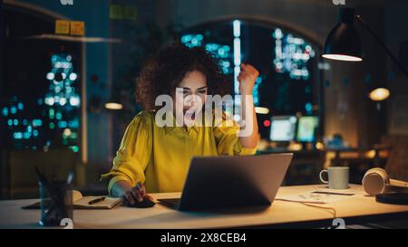 Expressive Middle Eastern Female Sitting Working on Laptop Computer in the Evening. Project Manager Receiving Positive News About Her Business Submission. Excited Specialist is Happy and Celebrating. Stock Photo