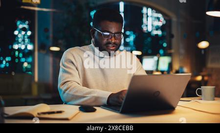 Slightly Agitated Male Employee Working on Laptop Computer in Office During Evening Time. Diverse Black African Specialist Browsing Internet, Researching Financial Data, Answering to a Work Email. Stock Photo