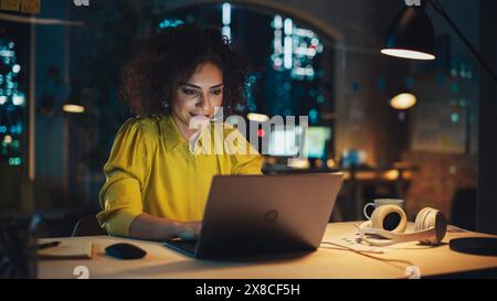 Creative Multiethnic Female Working on Laptop Computer in a Company Office. Happy Project Manager Browsing Internet, Writing Tasks, Developing a Marketing Strategy for Clients. Stock Photo