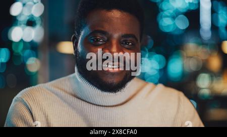 Close Up Portrait of a Handsome Black Man Wearing Stylish White Turtleneck, Looking at Camera and Expressing a Perfect Smile. Happy Attractive Manager at Work in Office in the Evening. Stock Photo