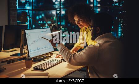 Team of Two Young Entrepreneurs Talking, Discussing Growth Strategy at a Desk with Computer. Stylish Businesspeople Work on an Investment and Marketing Report in a Creative Agency in the Evening. Stock Photo