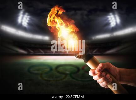 Hand holding lit Olympic torch with stadium behind with lights and Olympic rings drawn on the grass. Front view. Stock Photo