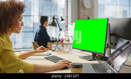 Portrait of a Young Creative Biracial Woman Working on a Computer with Green Screen Display in Modern Office. Female Creative Software Developer Using Chroma Key Display on Monitor. Stock Photo