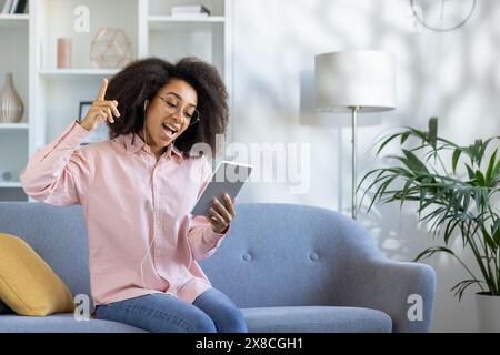 In a contemporary living space, a joyful young woman with curly hair and glasses, dressed in a pink shirt, relaxes on a blue couch while listening to music on a tablet, exuding happiness Stock Photo