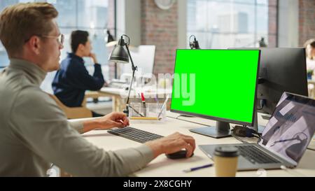 Portrait of Young Caucasian Man Working on Laptop and Green Screen Computer During Day in Modern Bright Office. Creative Software Developer Using Chroma Key Display PC Stock Photo
