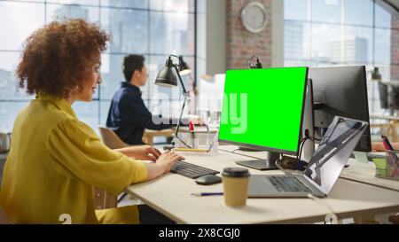Employees Smiling While Working on Computers in Busy Modern Bright Office. Biracial Woman Designing Using Green Screen. Male Employee Working in the Background. Stock Photo