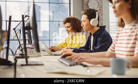 Multiethnic Group of Creative Colleagues Working on Computers in a Modern Bright Office. A Biracial Woman Smiles Happily After Successfully Finishing a Work Project. Asian Man Using Headphones Stock Photo