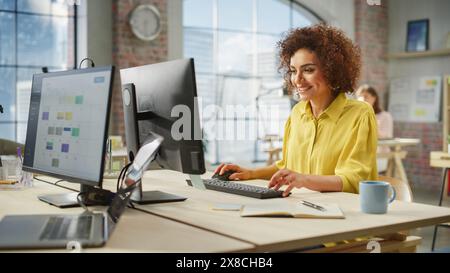 Portrait of Biracial Creative Young Woman Working on a Computer in Bright Busy Office. Female Recruiter Smiling Happily When Receiving Positive Feedback. Employee Working in Background. Stock Photo