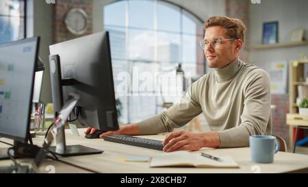 Portrait of Caucasian Creative Young Man Working on Computer During Day in Modern Office. Supervisor Smiling While Answering Email Questions From Team Memebers. Stock Photo