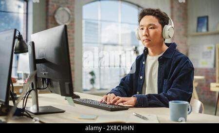 Portrait of Asian Male Employee Working on Computer in a Modern Office During the Day. Customer Support Agent Answering Requests While Using Headphones. Stock Photo