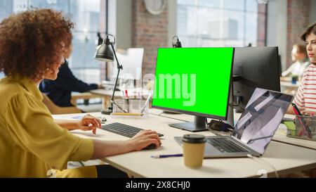 Biracial Woman Smiles While Using Green Screen Display on a Computer in a Busy Bright Office. Multiethnic Group of Creative Colleagues Working on Computers in the Background. Stock Photo