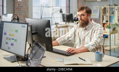Portrait of Creative Bearded White Man with Glasses Focused While Using Computer in Bright Spacious Office. Marketing Manager Building a New Strategy. Stock Photo