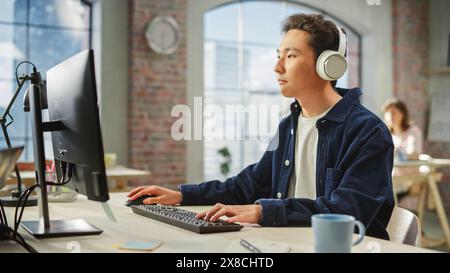 Portrait of Asian Creative Man Using Computer and Headphones in Bright Modern Office. Male Data Analyst Developing Database While Listening to Music. Stock Photo