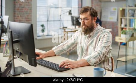 Portrait of Young Caucasian Bearded Man with Glasses Focused While Sitting and Working on Computer in a Bright Spacious Office. Male Legal Consultant Typing Notes on Keyboard. Stock Photo