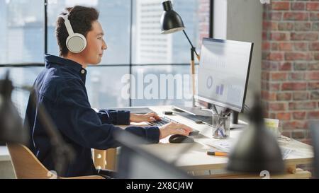 Profile View of a Portrait of an Asian Young Man Working on a Computer and Using Headphones in a Modern Office. Data Analyst Reviewing Charts and Statistics. Stock Photo
