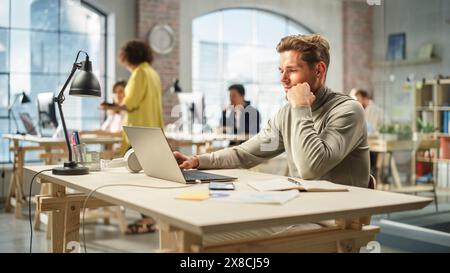 Portrait of Caucasian Productive Young Man Working on Laptop in an Active Office. White Businessman Developing an E-commerce Strategy. Busy Colleagues Working in the Background. Stock Photo