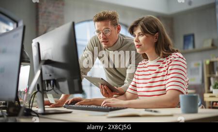 Two Young Creative Colleagues Discussing Work Project and pointing at the computer screen. Caucasian Male Supervisor Uses a Tablet and Consults Female Team Leader. Stock Photo