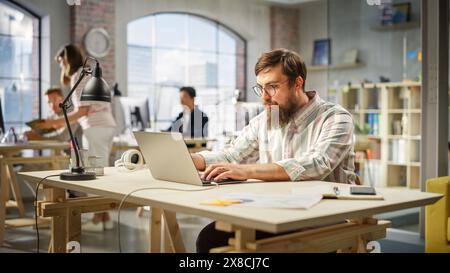 Portrait of Motivated Caucasian Young Man With Beard Working on a Laptop in Office. Male Director Preparing a Marketing Project. Diverse Team Working on Computers in the Background. Stock Photo