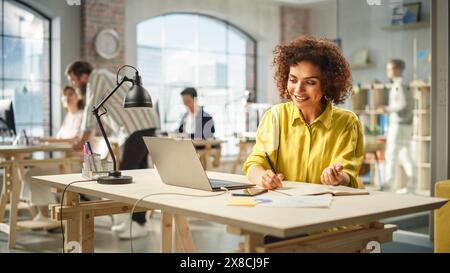 Portrait of Young Biracial Woman Taking Notes at Her Desk While Looking for References on the Laptop. Busy Employees Are Collaborating in the Background. Stock Photo